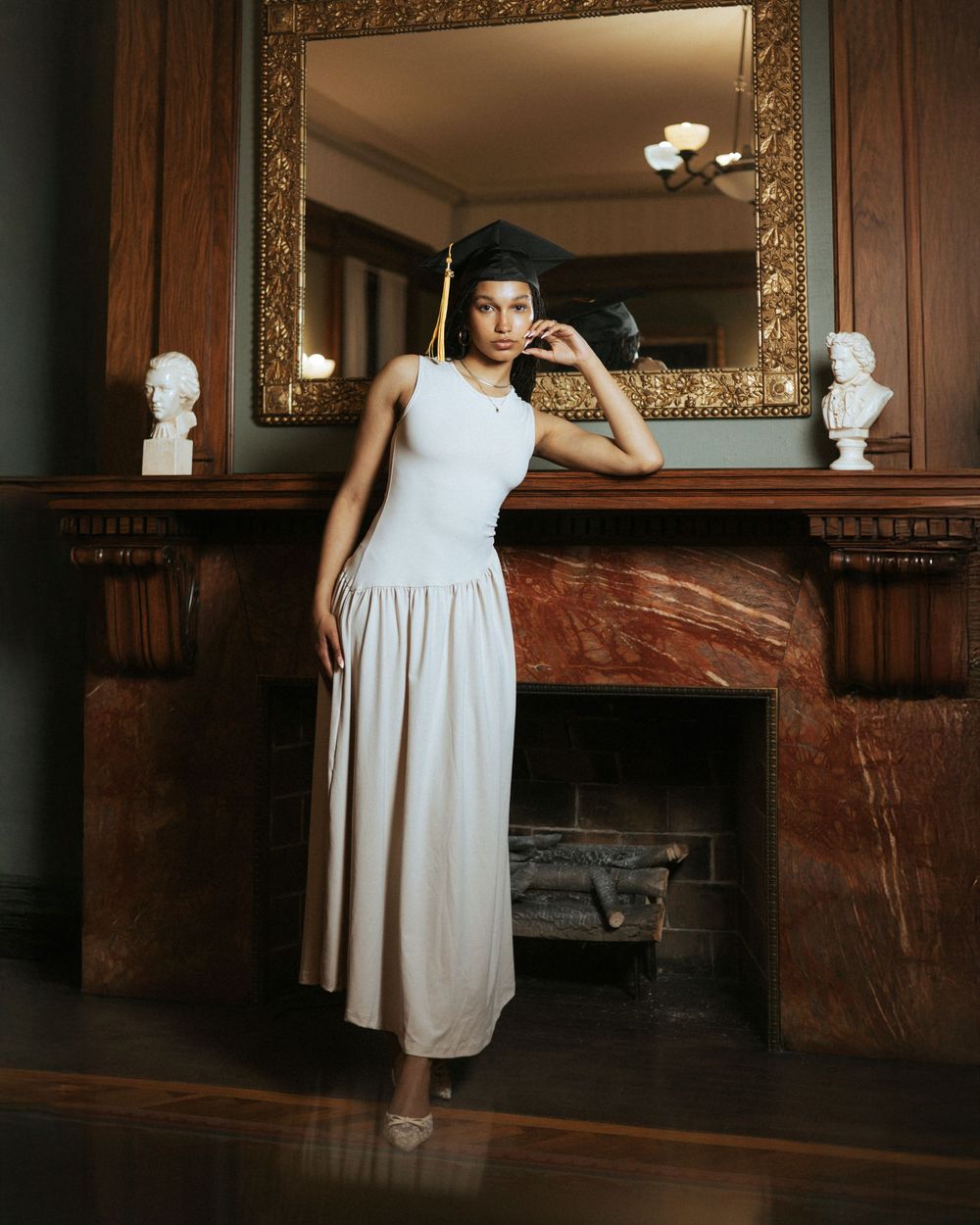 Stylish figure in a long white dress poses elegantly beside an ornate marble fireplace with decorative busts and mirror.