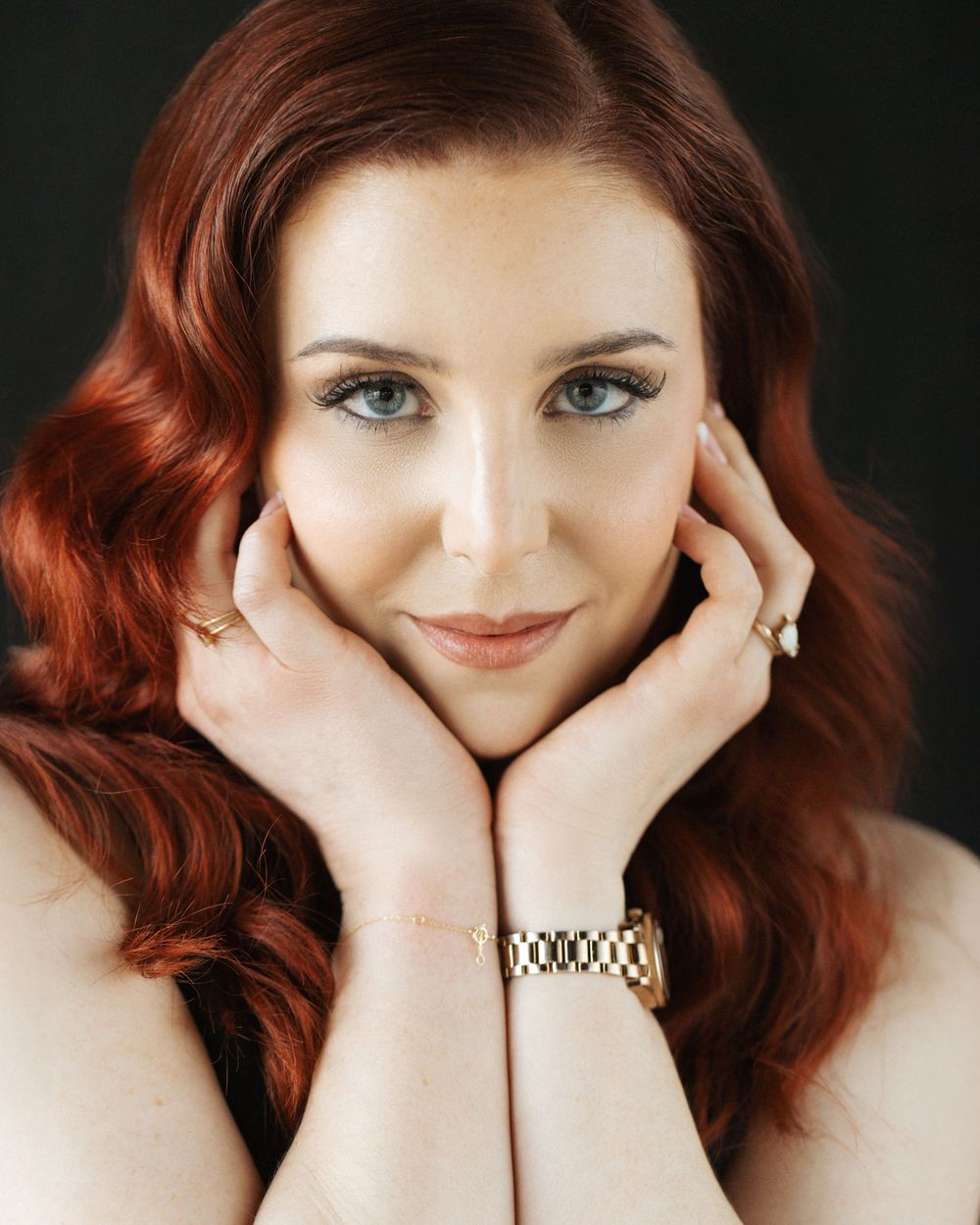 Portrait shot featuring vibrant red wavy hair, blue eyes, and gold jewelry against a dark background.