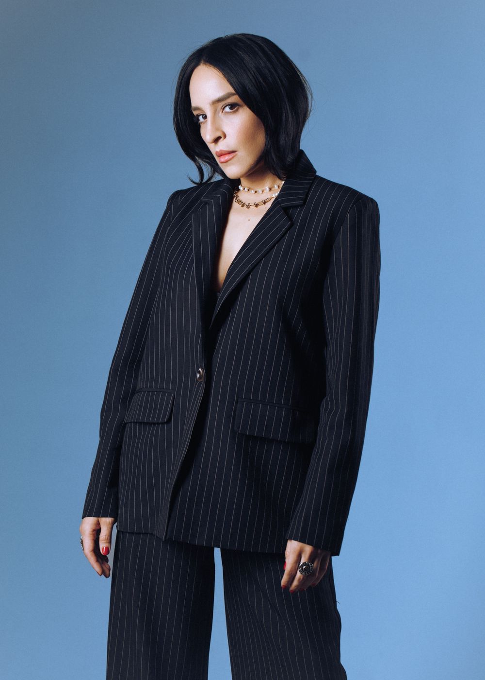 A person wearing a black pinstripe suit and gold necklace poses against a blue studio backdrop.