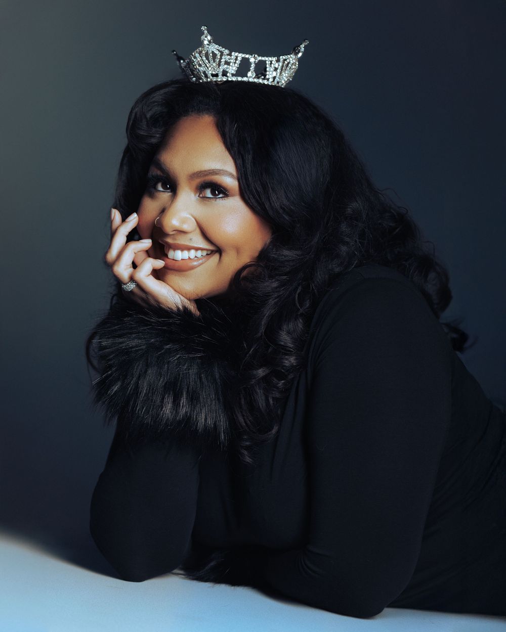 Woman wearing a sparkling tiara crown poses elegantly in a black outfit, smiling confidently against a dark studio background.