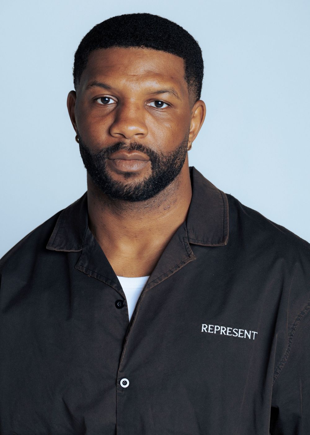 Man wearing a dark Represent branded shirt against a light blue background, with short hair and beard.