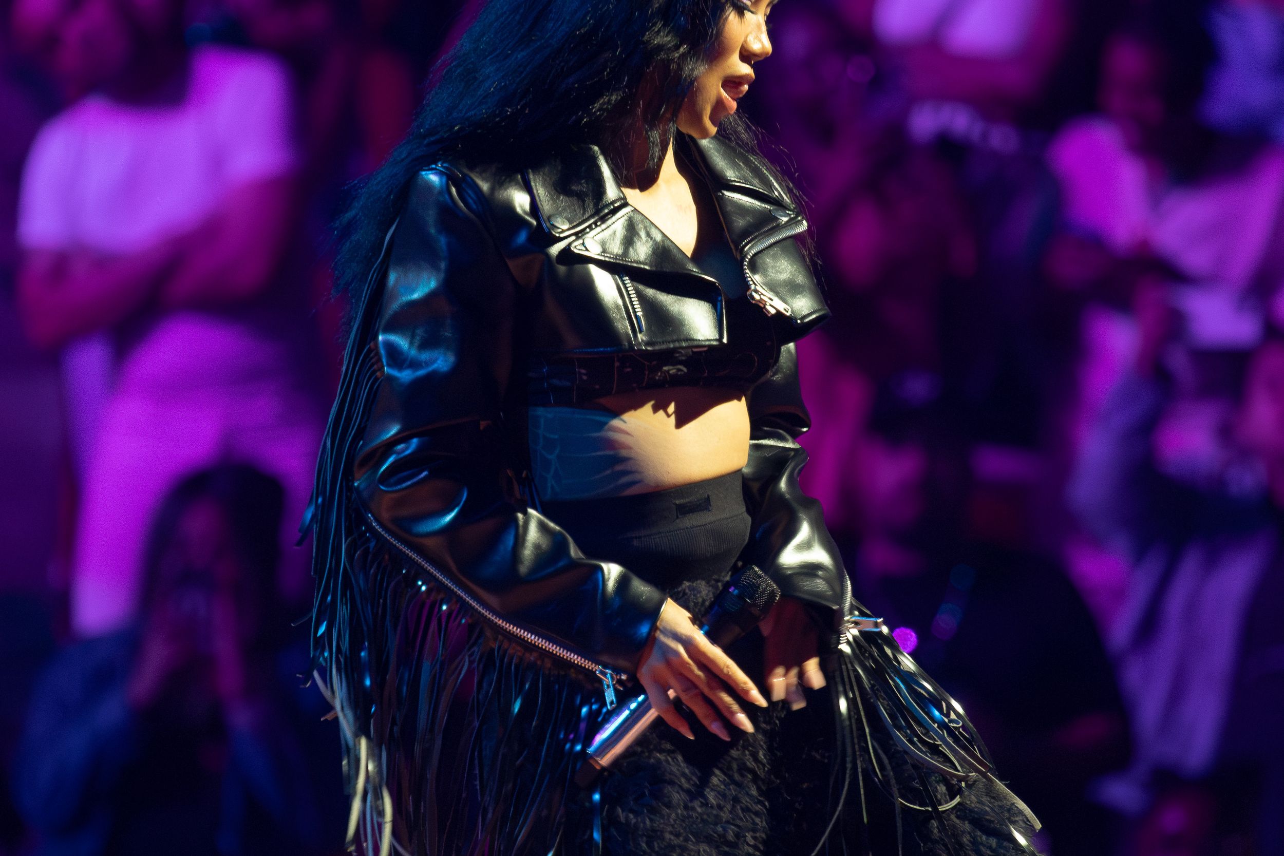 Performer in black leather outfit holding microphone on purple-lit stage.