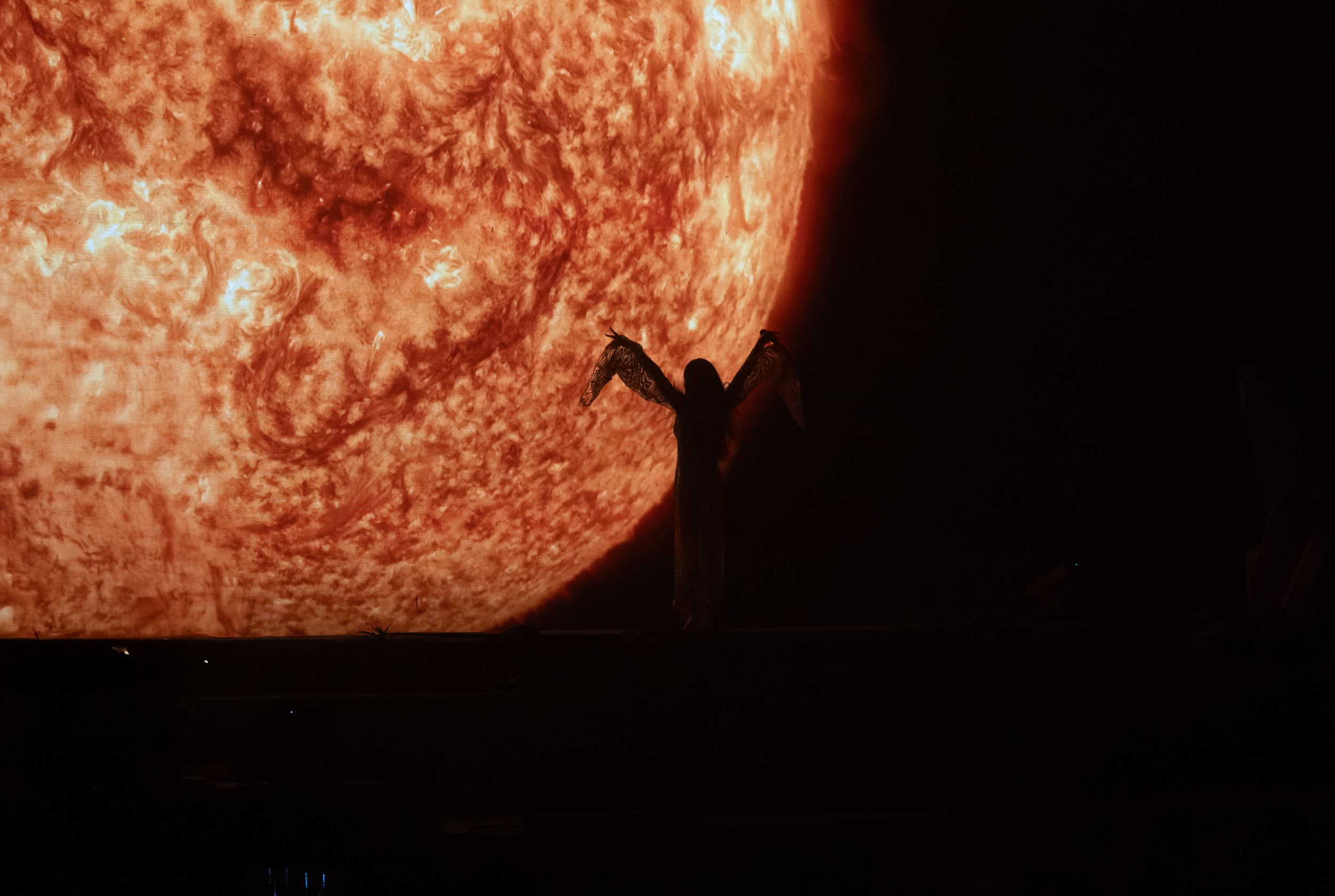 Silhouette of person reaching toward large glowing orange sun against black background.