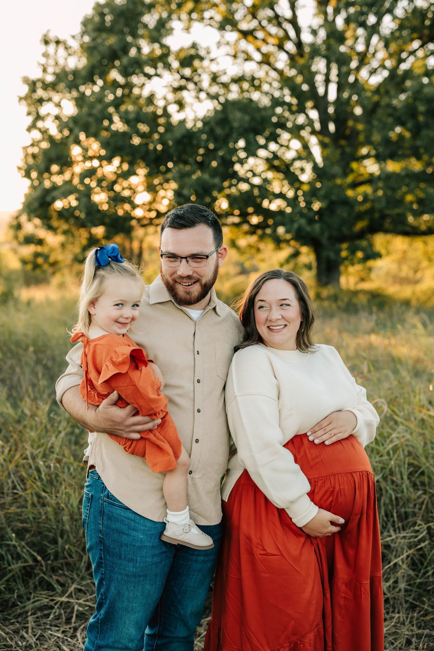 A family stands in a sunlit field with golden trees, wearing coordinated orange and white outfits during sunset.