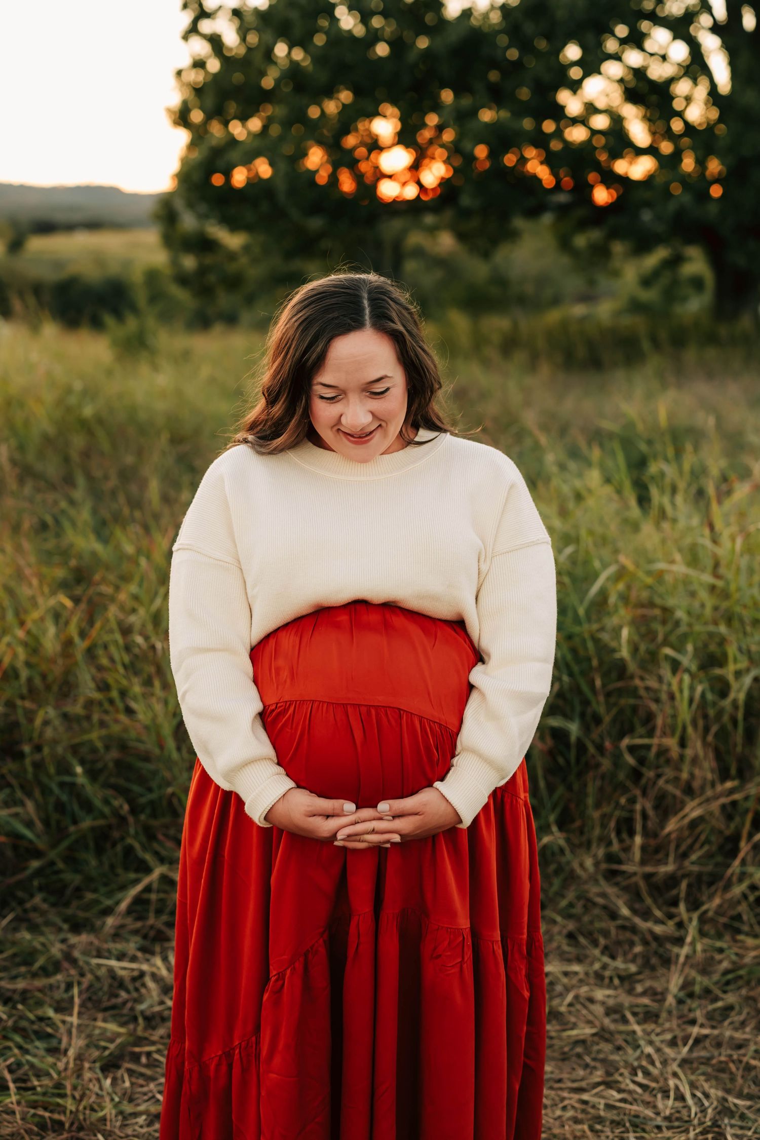Expectant mother in cream sweater and flowing red dress cradles baby bump during golden hour sunset in field.