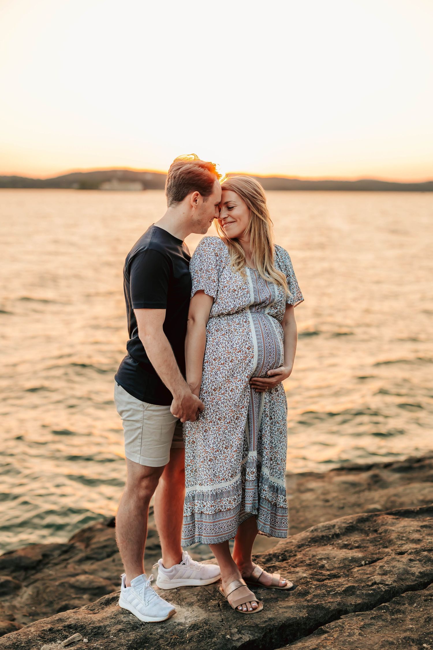 A loving couple stands together on rocky shore at sunset while expecting a baby.