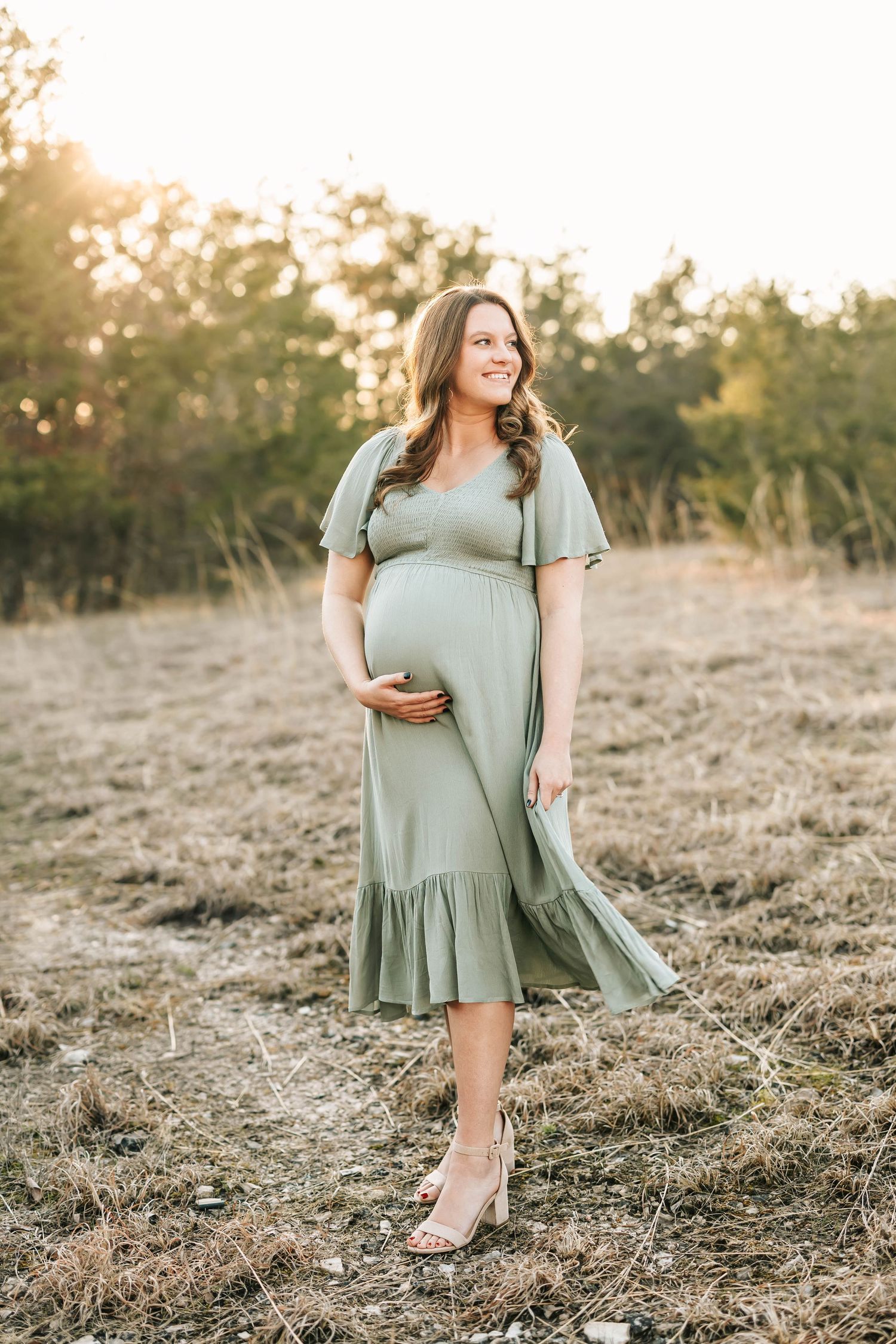 Expectant person wearing a sage green midi dress poses in a sunlit field at golden hour, cradling baby bump.