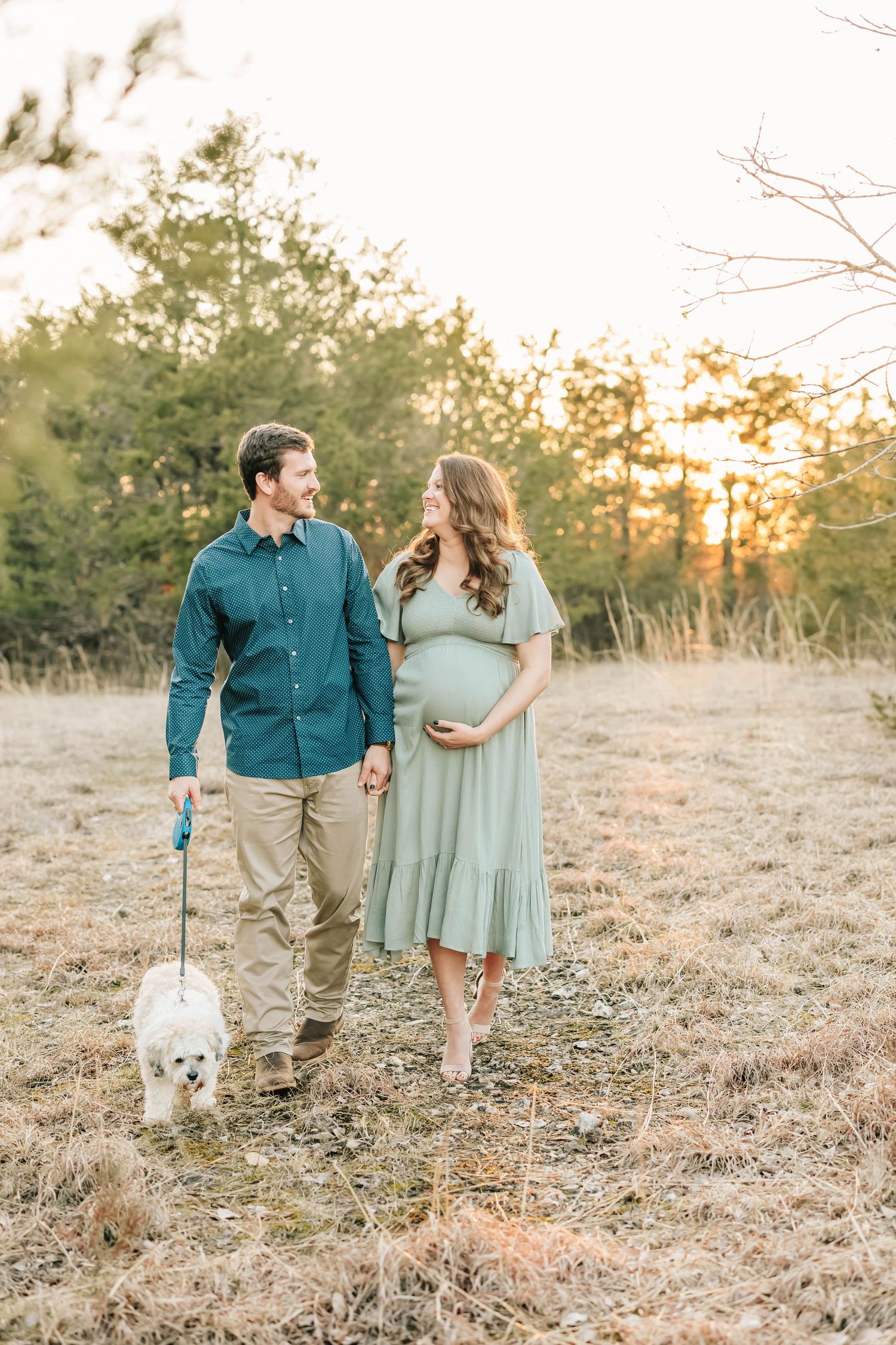 Happy couple walking their small white dog in a field at sunset while expecting their first child.