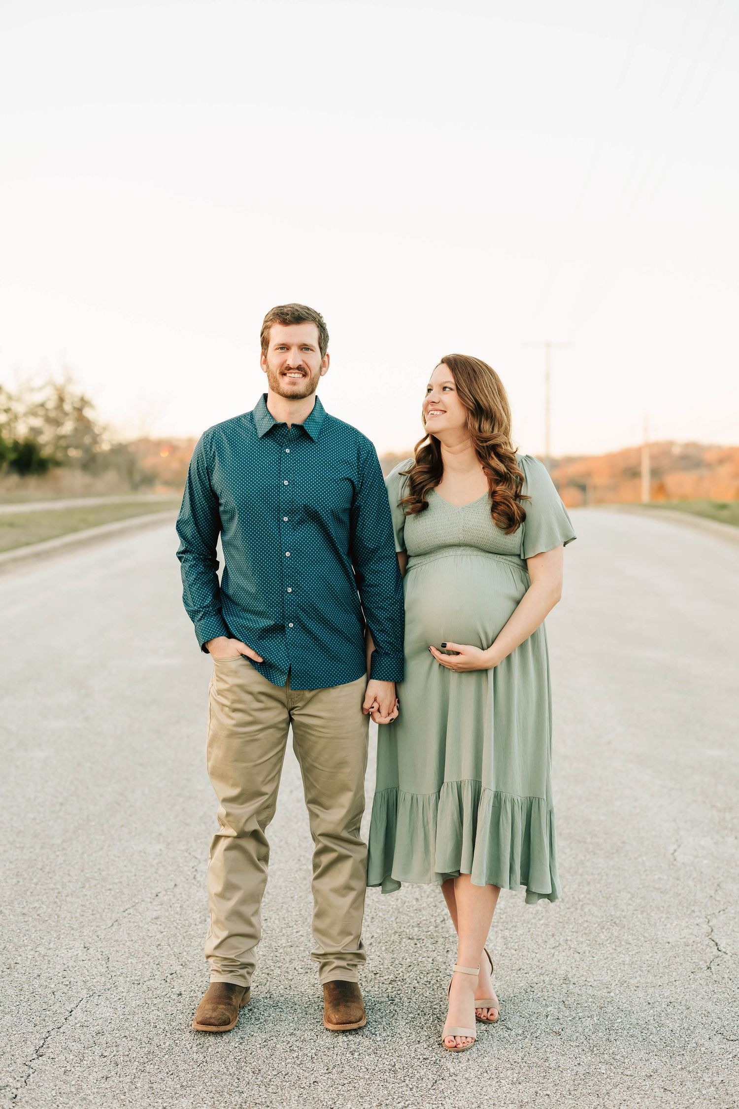 A couple stands together on a rural road during a maternity photo session at sunset.