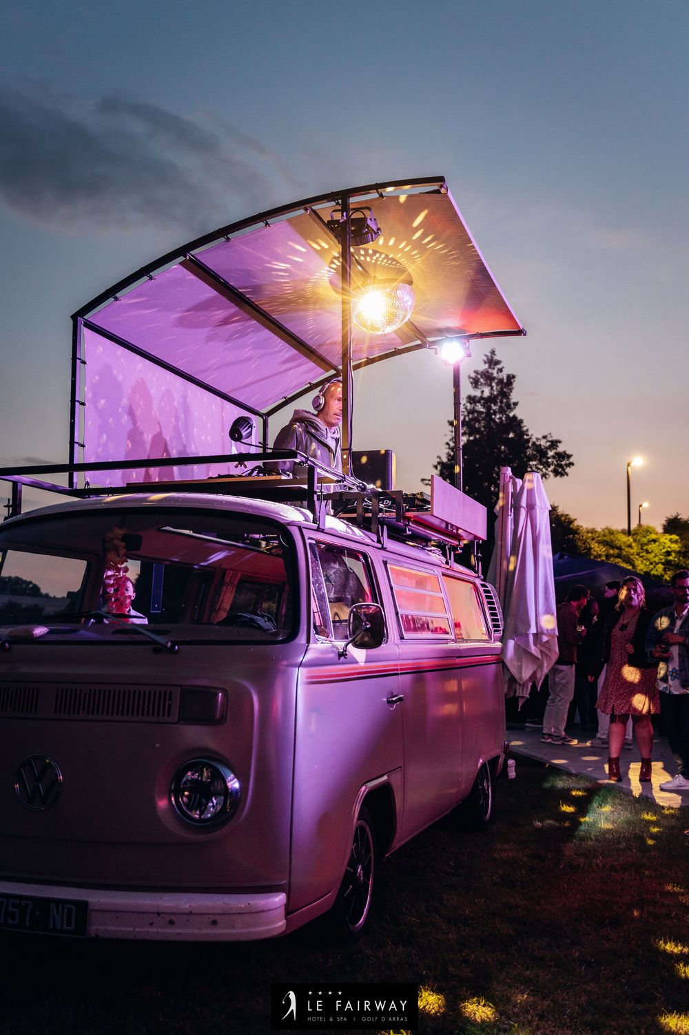Vintage pink Volkswagen van converted into mobile DJ booth with light display at dusk outdoor event.