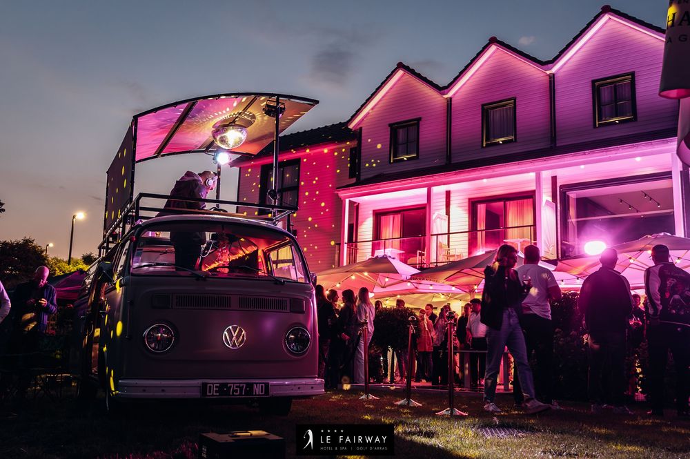 Vintage VW van DJ booth at Le Fairway evening outdoor party with pink-lit hotel building backdrop.