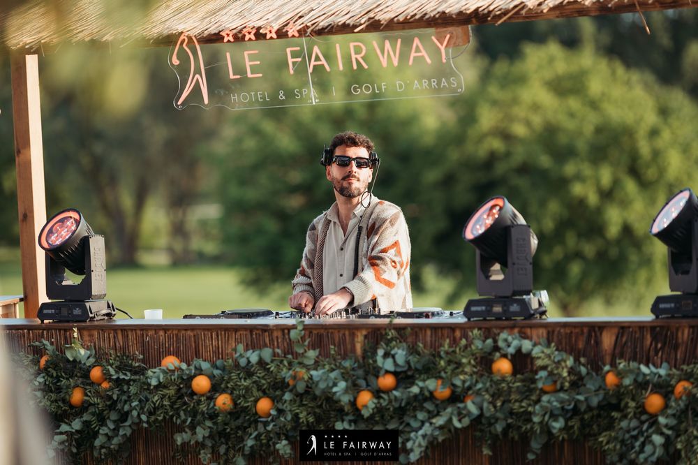 DJ performing at Le Fairway Hotel Golf outdoor event under thatched roof with stage lighting.