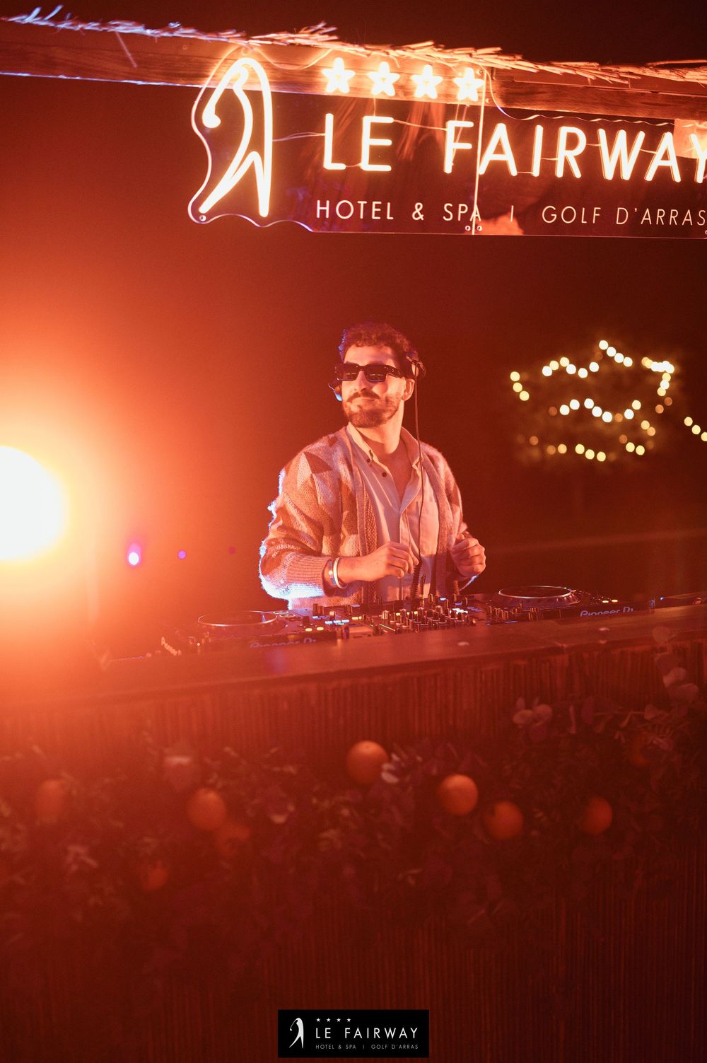 DJ performing at night under Le Fairway Hotel Spa Golf d'Arras illuminated sign with warm orange lighting.