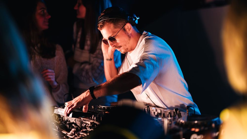 DJ wearing sunglasses performing at a nightclub event, focused on mixing equipment with crowd in background.