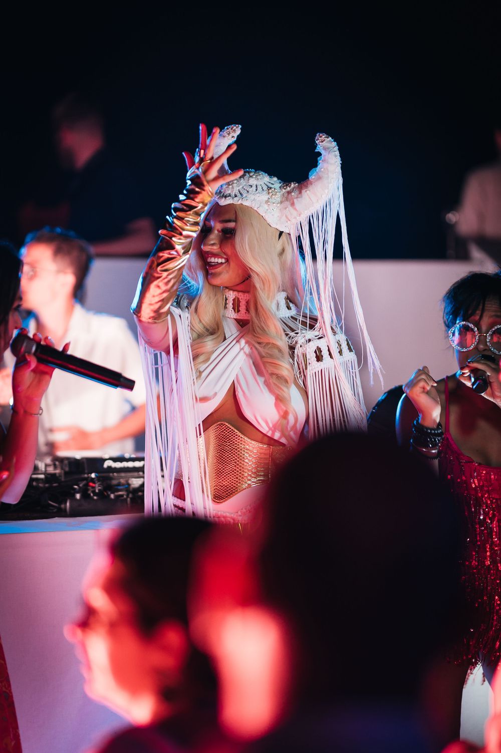 Performer in white fringe headdress and gold corset dancing at a nightclub event under colorful lights.