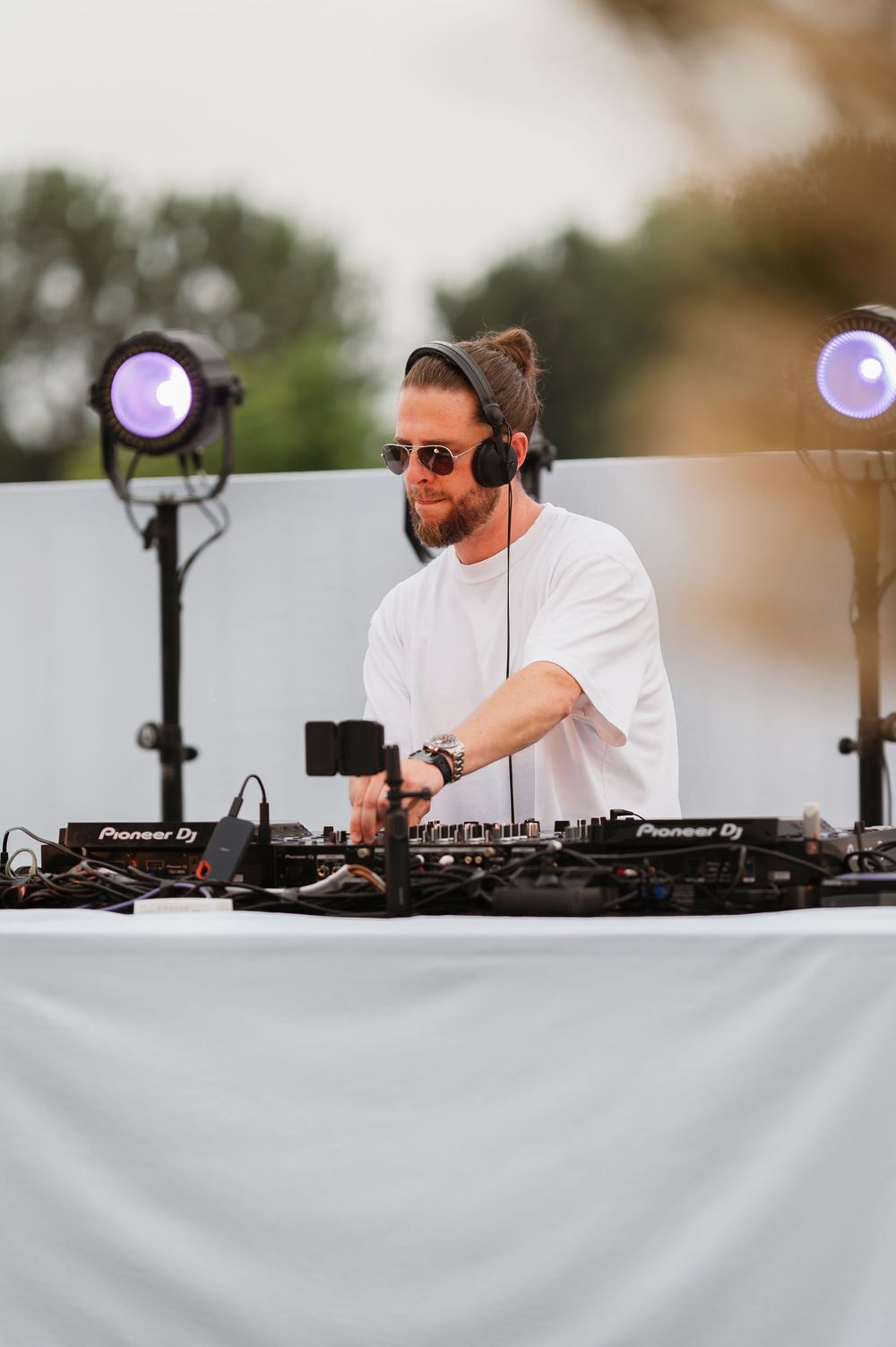 A bearded DJ wearing sunglasses and headphones performs at an outdoor Pioneer DJ setup with stage lights.