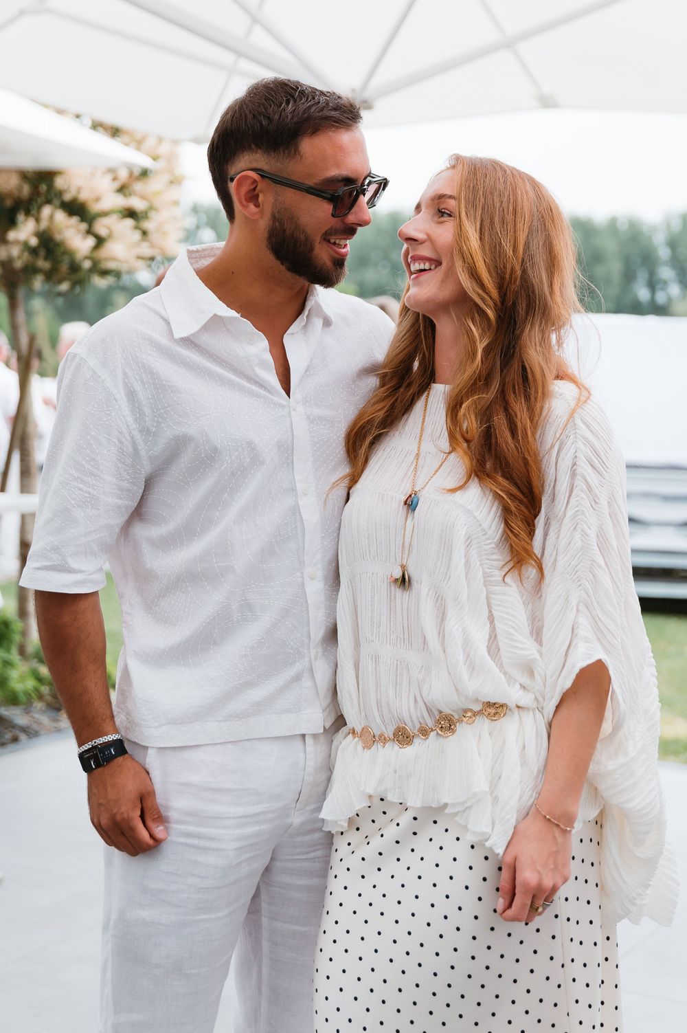 A smiling couple dressed in white outfits poses together outdoors, the woman in a polka dot skirt and the man in sunglasses.
