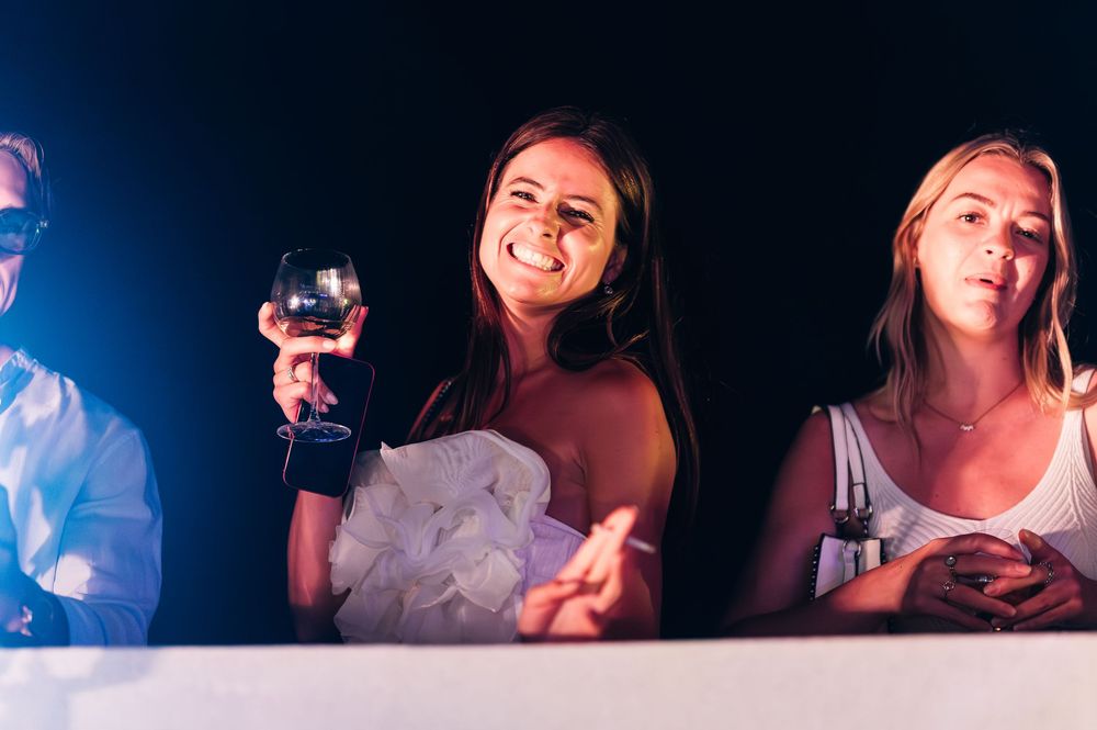 Two young women smiling at a party, one holding a wine glass, illuminated by colorful lights at a nighttime event.