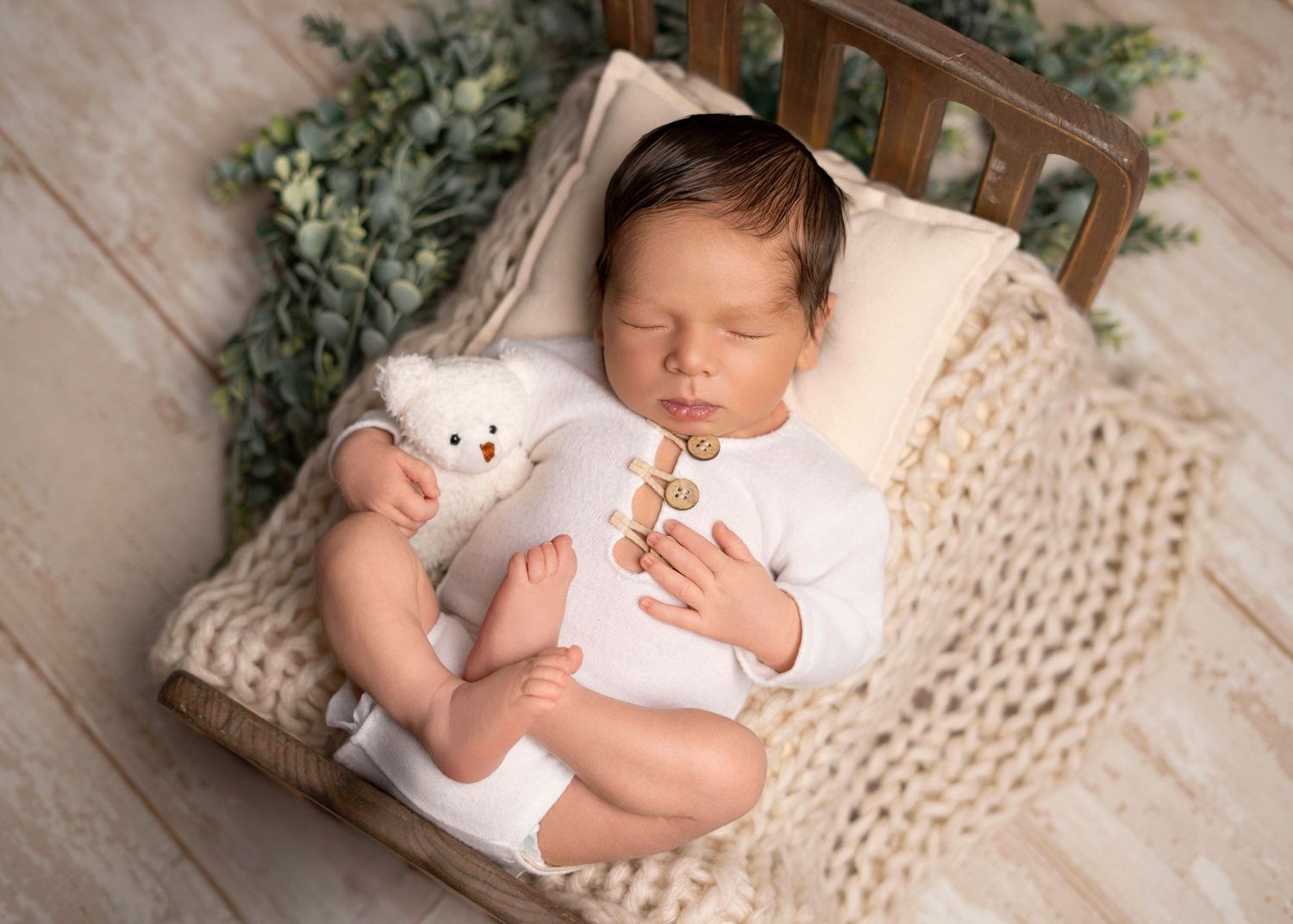 Newborn photographer Covina CA baby boy in white outfit posed in cozy basket with soft greenery sleeping in neutral studio setup with teddy