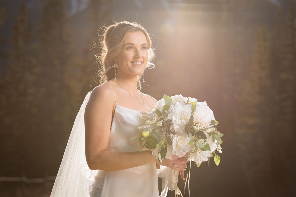 Radiant bride in white gown holding bouquet of blush roses, bathed in golden sunlight with veil flowing.