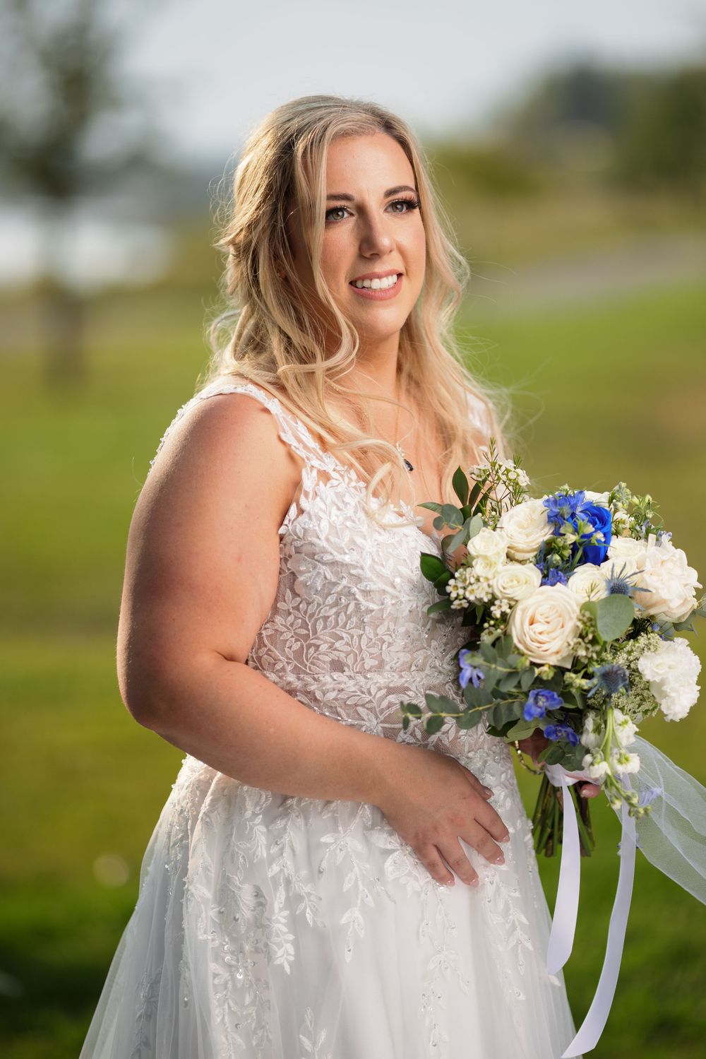 Blonde bride in lace wedding dress holds blue and white bouquet while smiling outdoors on green lawn.