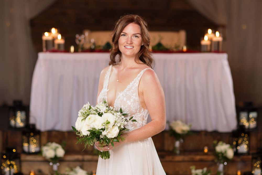 Smiling bride in white lace gown holding bouquet of white flowers, standing before candlelit reception table.