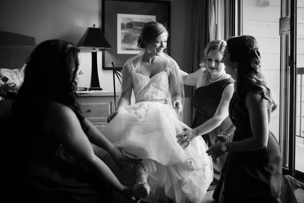 Black and white photo of a bride in a white gown being helped by bridesmaids in a warmly lit room.