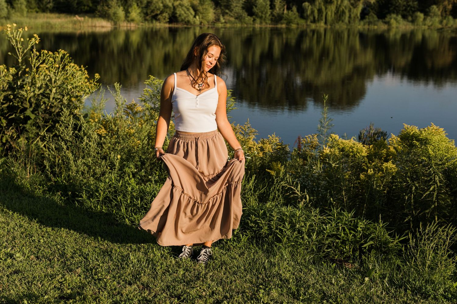 Woman in flowing beige dress twirling by a serene lake at golden hour.