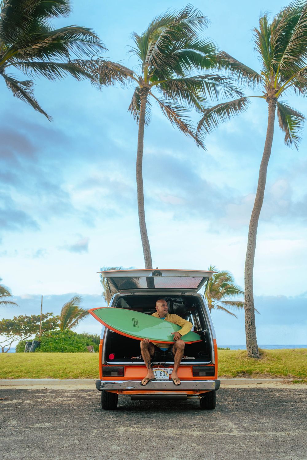 Surfer sitting in open trunk of orange van holding green surfboard with palm trees and ocean in background.