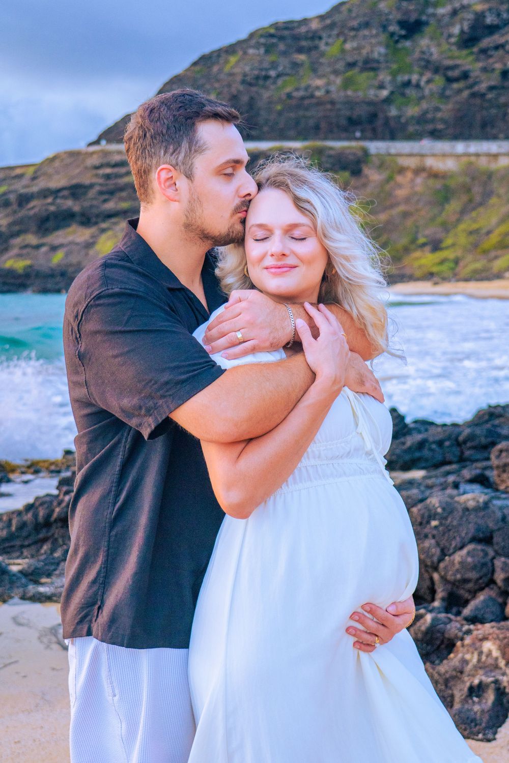 Man tenderly kisses and embraces pregnant woman in white dress on rocky Hawaiian beach at sunset.