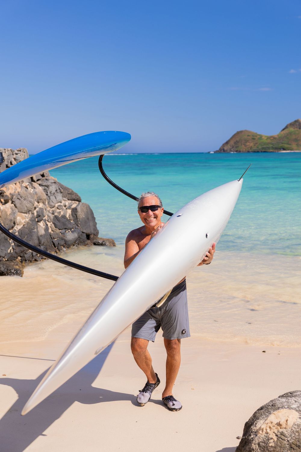 Man holding a white kayak on a tropical beach with turquoise water and blue sky in the background.