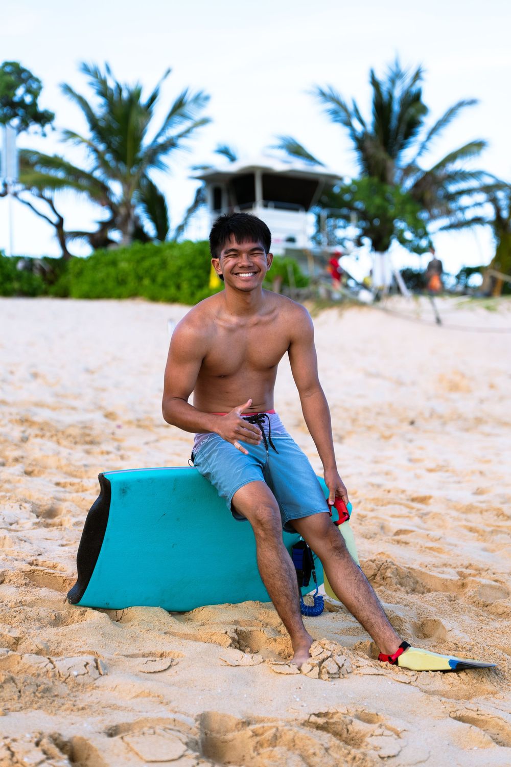 Smiling young man sitting on a bodyboard on a sandy beach with palm trees and a lifeguard tower in the background.
