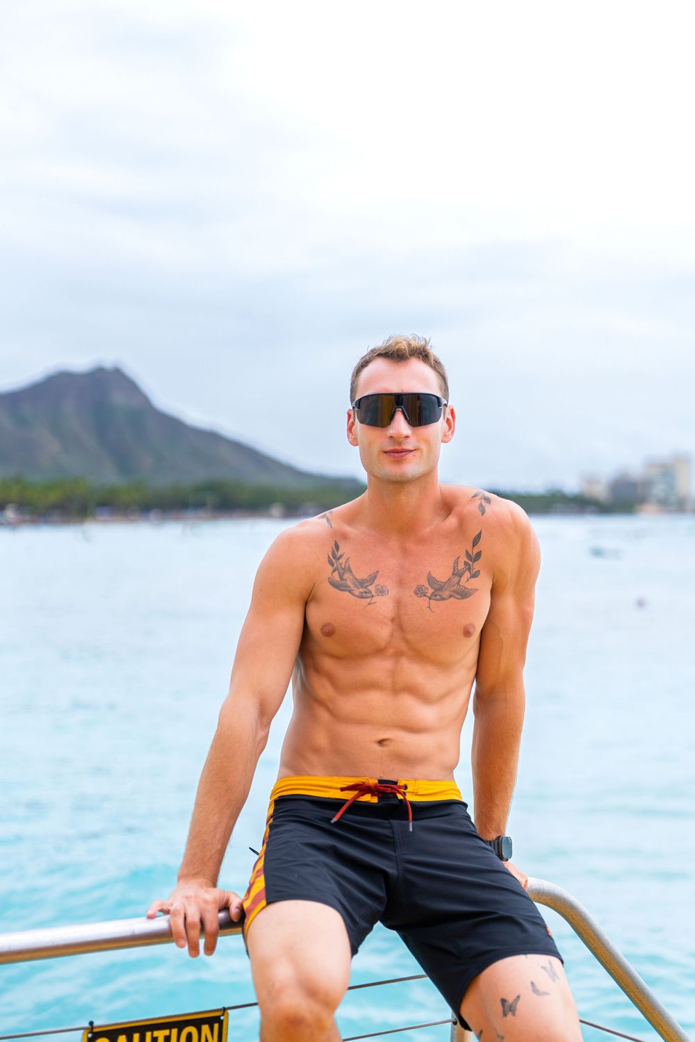 Shirtless tattooed man in sunglasses and swim shorts on a boat with Diamond Head crater in background.
