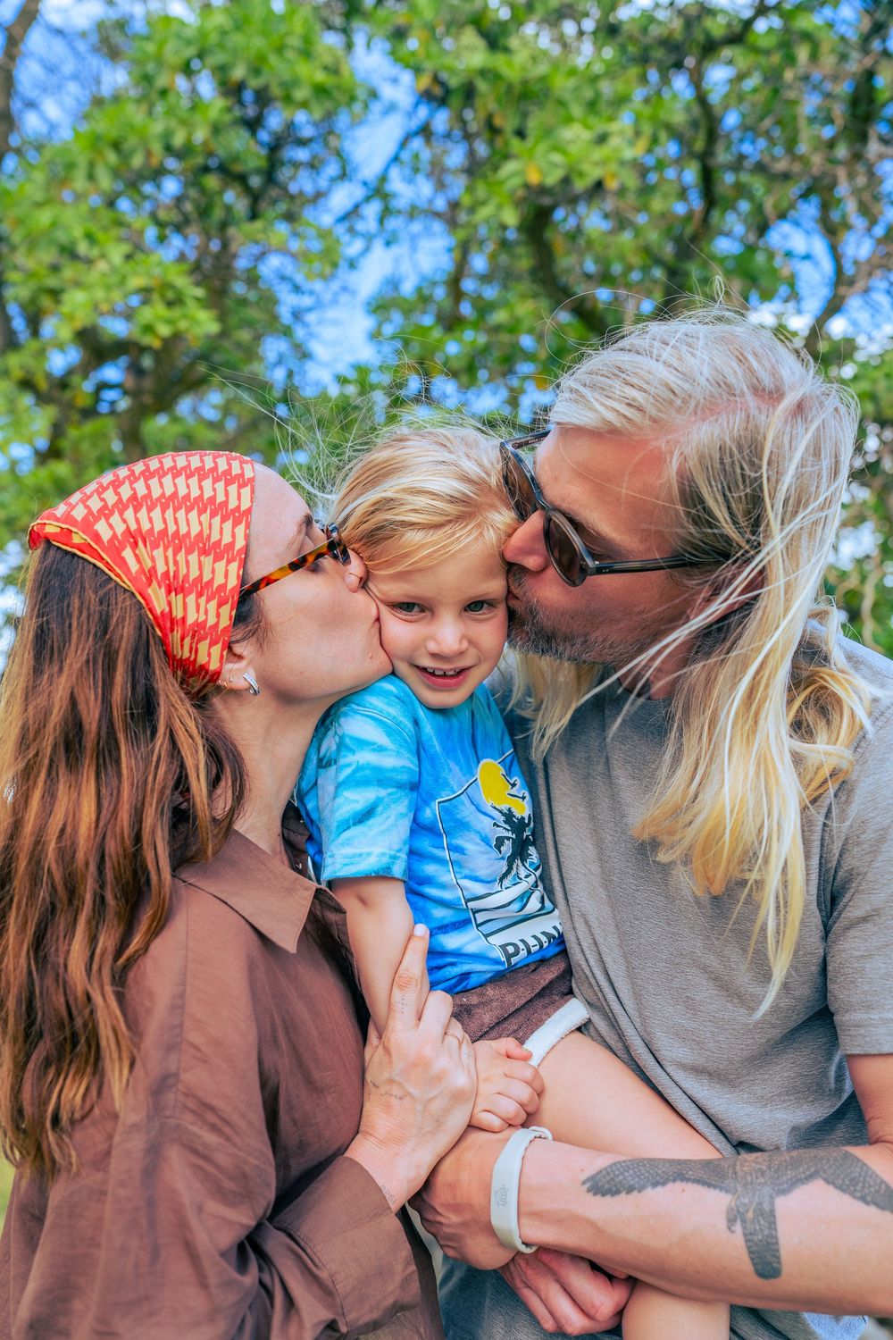 Two parents kissing their smiling blond toddler son outdoors under lush green trees.