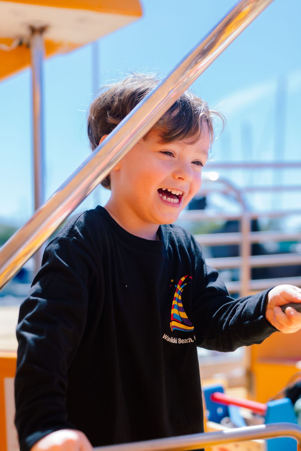 Joyful young boy in a black Waikiki Beach shirt laughing while playing on a sunny outdoor structure.