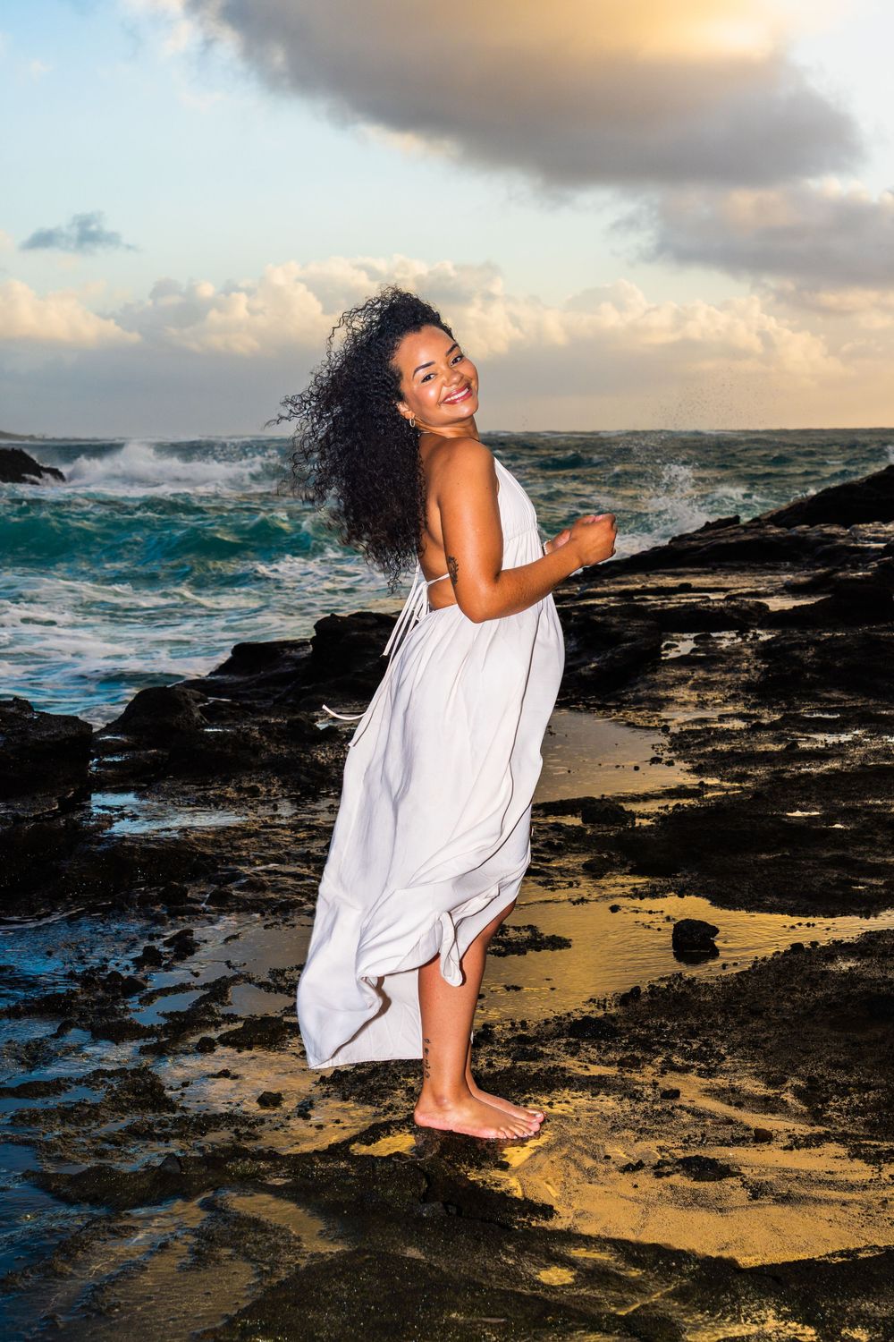 Smiling woman in white dress standing on rocky beach at sunset with ocean waves crashing behind her.