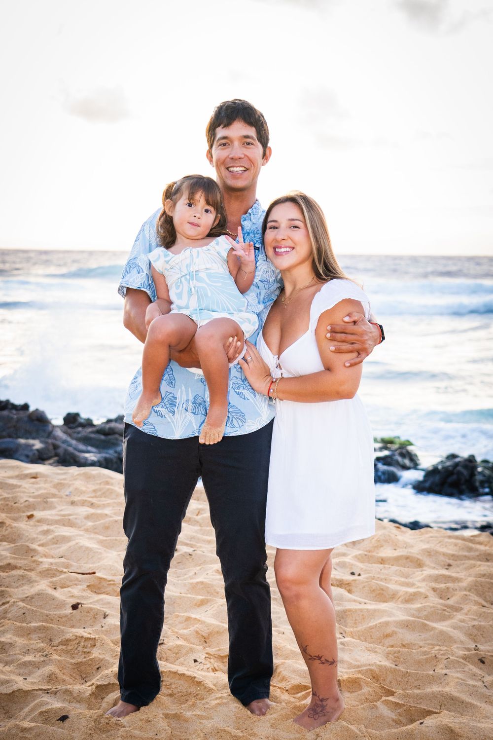 Happy family of three poses on sandy Hawaii beach at sunset with rocky shore and ocean waves.