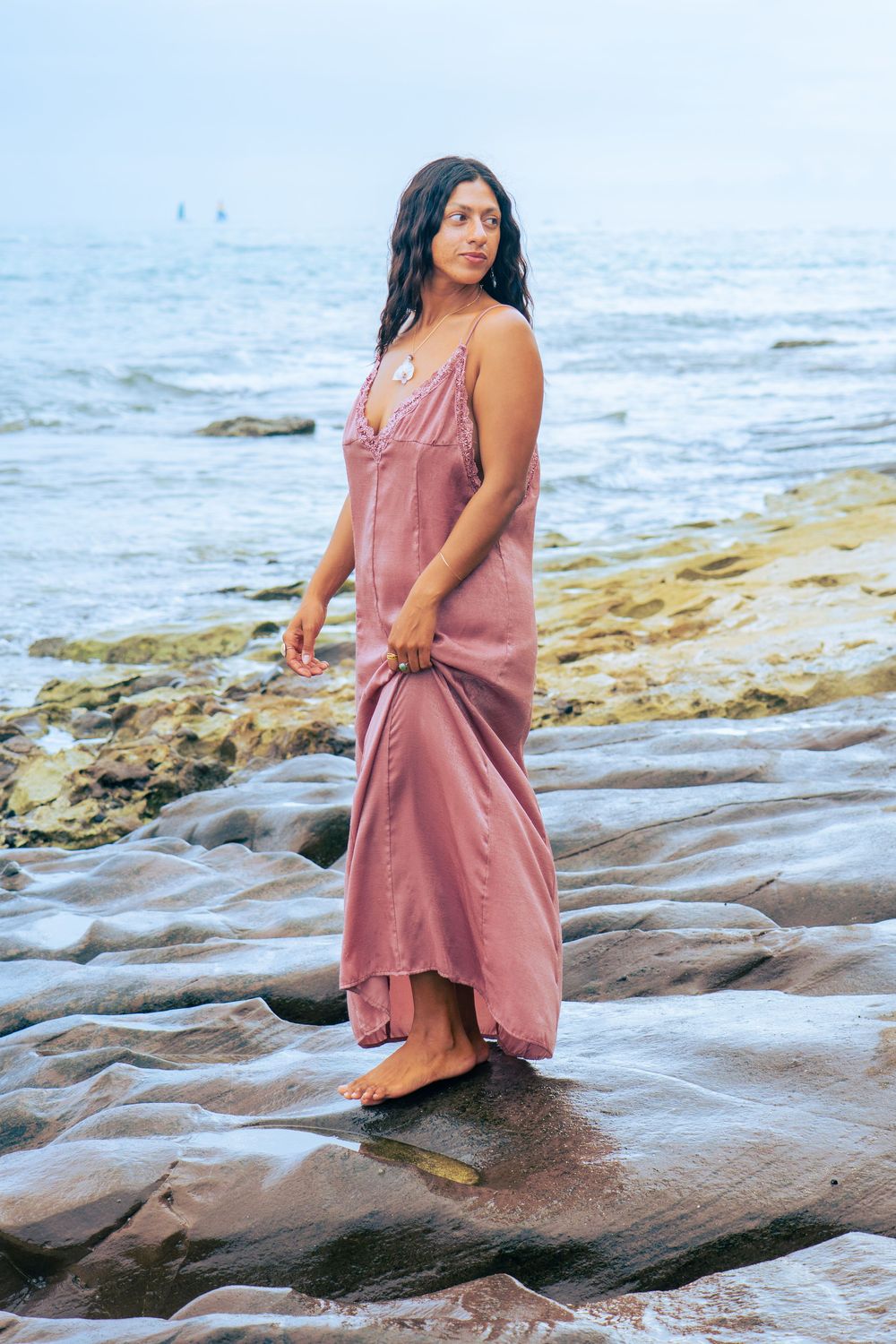 Woman in pink satin maxi dress standing on wet rocks by the ocean at dusk.