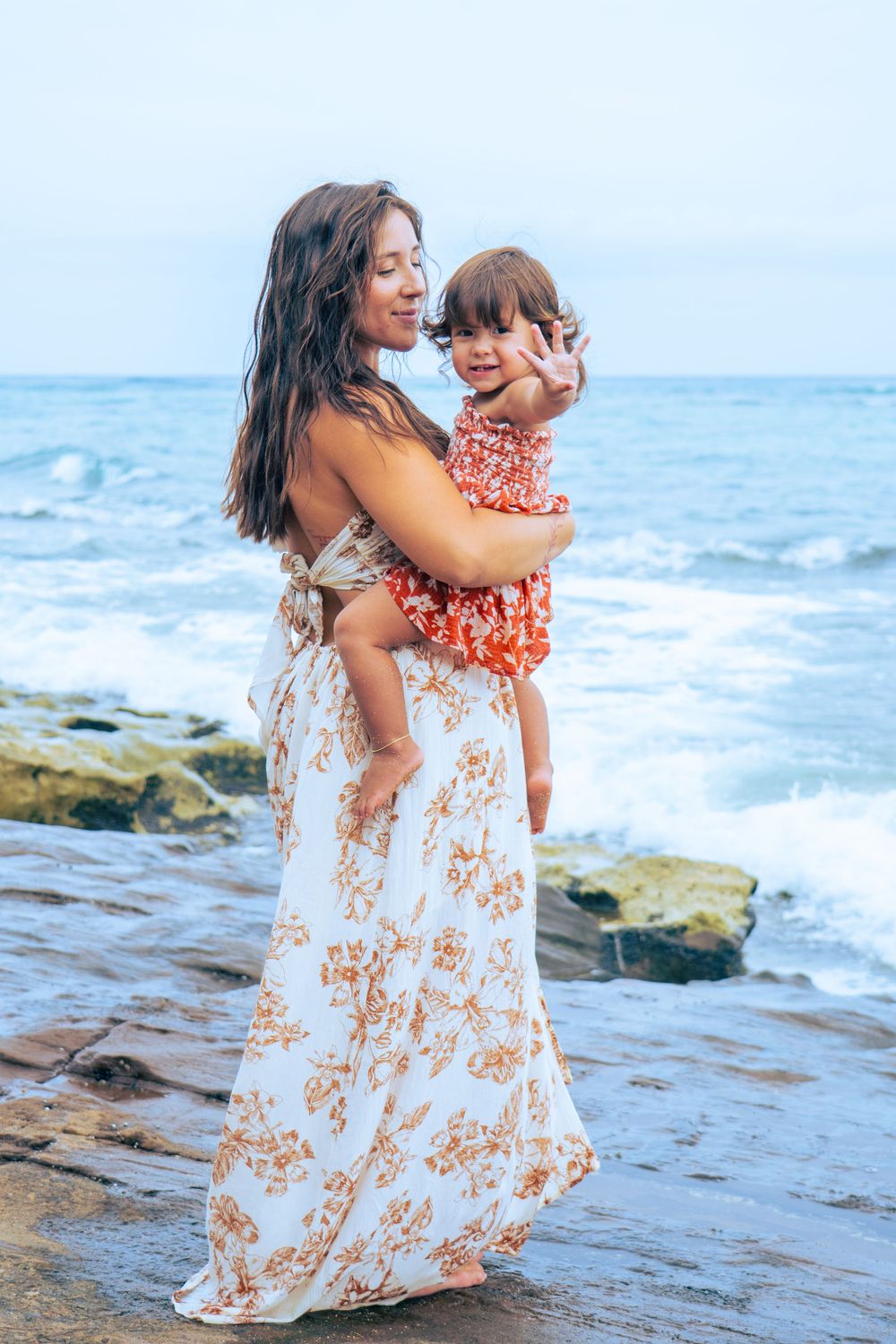 Mother in floral maxi dress holding young child on rocky shoreline with ocean waves behind them.