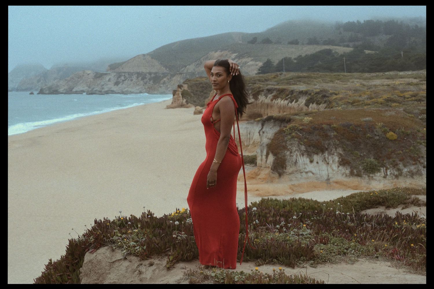 A person in a flowing red dress stands on a coastal cliff overlooking a misty beach and ocean landscape.
