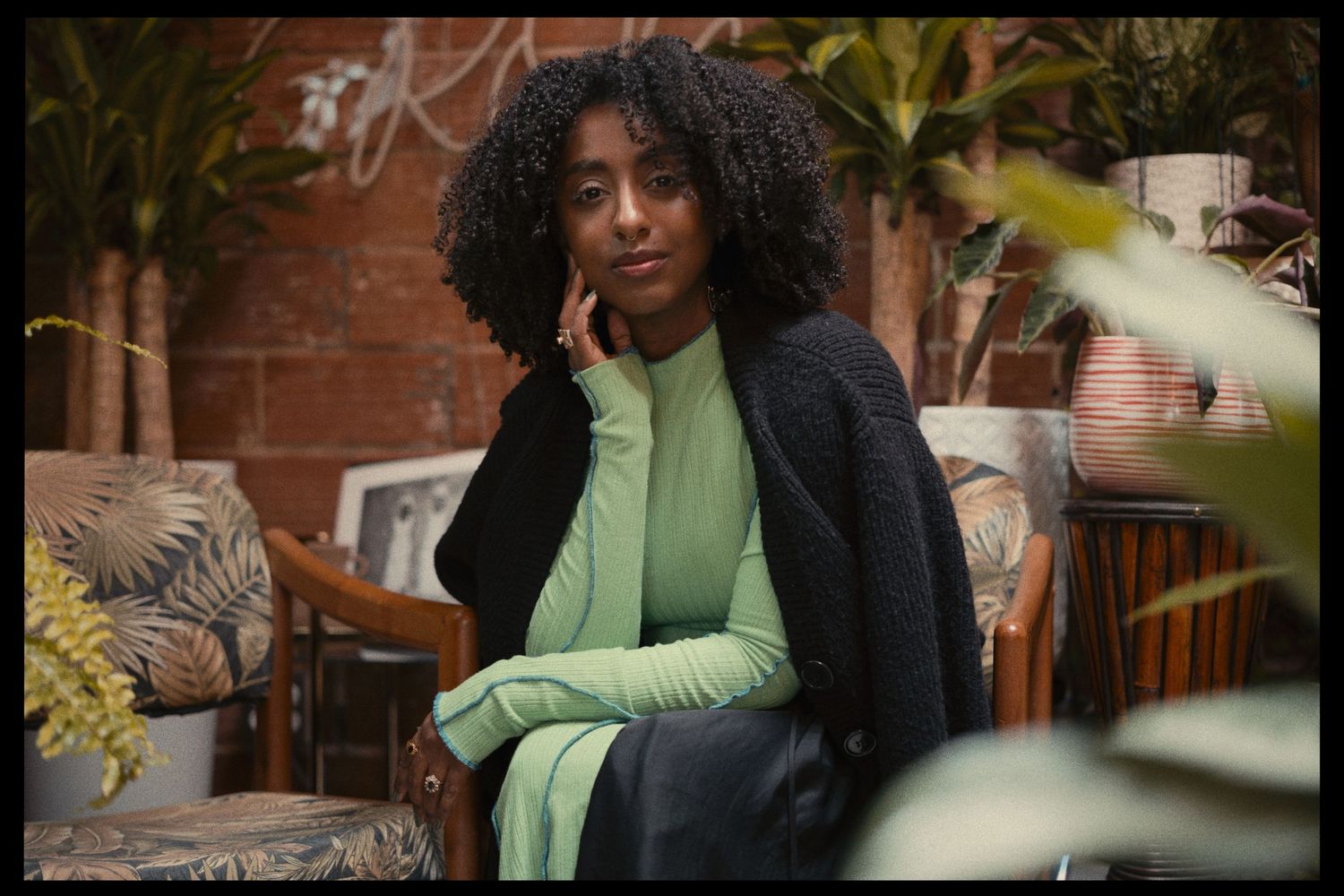 Young woman with curly hair in green turtleneck and navy cardigan sitting among lush plants and vintage furniture.