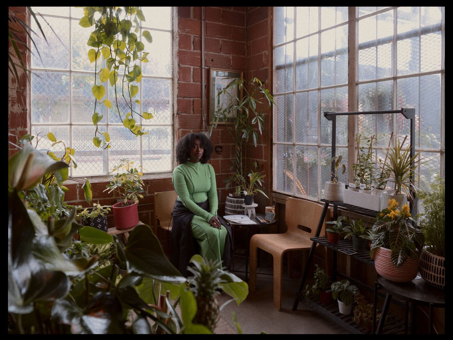 Woman in green outfit sits among lush indoor plants by large windows in a sunlit brick-walled room.