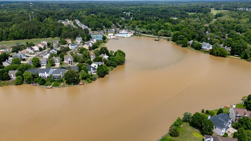 Aerial view of houses along a brown river after flooding with dense forest in the background.
