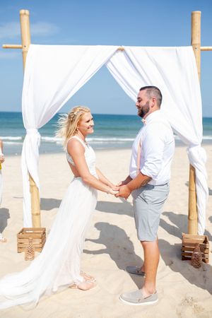 Beach wedding ceremony setup with white flowing curtains and wooden posts on sandy shore.
