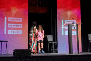 Stage presentation with red lighting and projection screens displaying policy information.