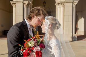 Romantic wedding portrait with red bouquet under stone archway.