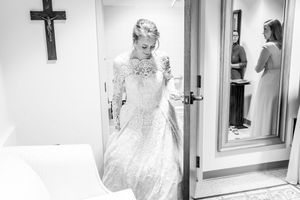 Black and white photo of bride in lace dress with cross on wall.