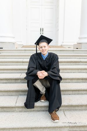 Young man in black graduation cap and gown sitting on stone steps, smiling at camera during graduation photo session.