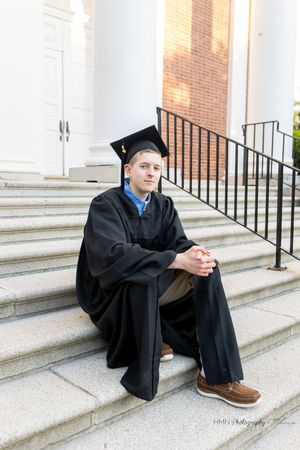 Young man in black graduation cap and gown sitting on stone steps outside a brick building.