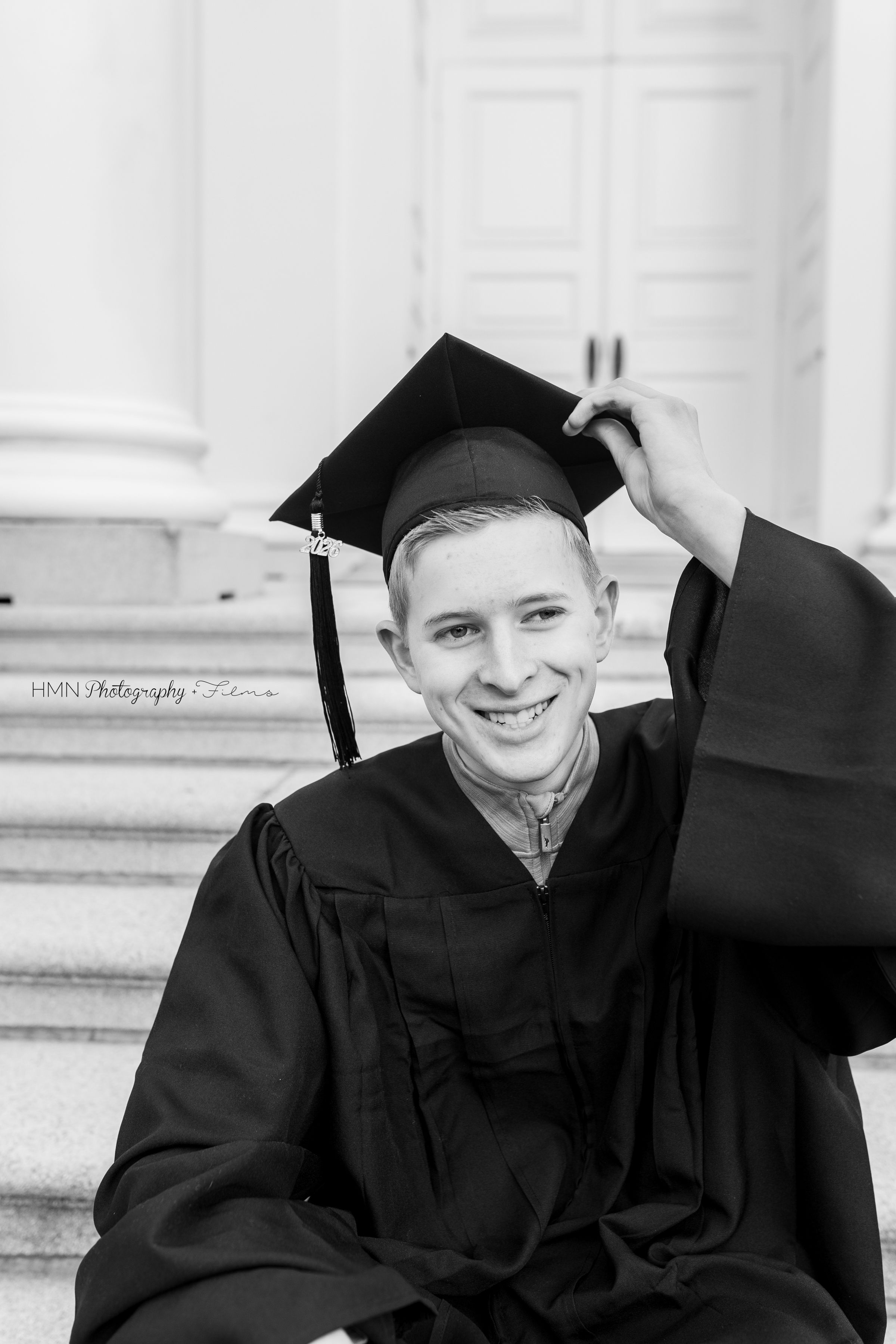 Smiling young man in black graduation cap and gown sitting on steps in a black and white photo.